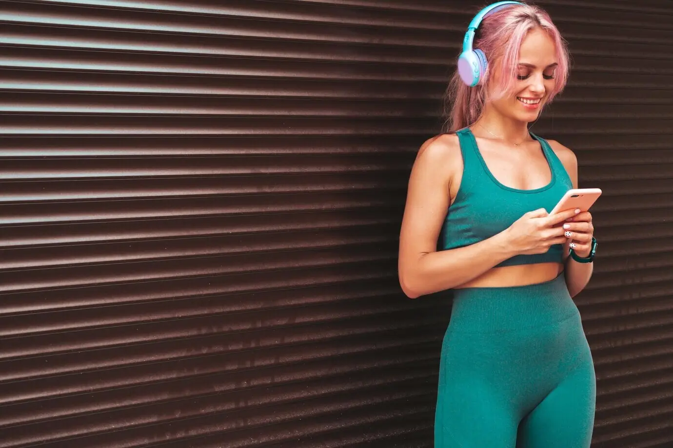 Mujer fitness sonriente con ropa deportiva verde y cabello rosa. Modelo joven y hermosa con cuerpo perfecto. En la calle, cerca de una pared con persiana enrollable, escuchando música con auriculares inalámbricos y usando aplicaciones.